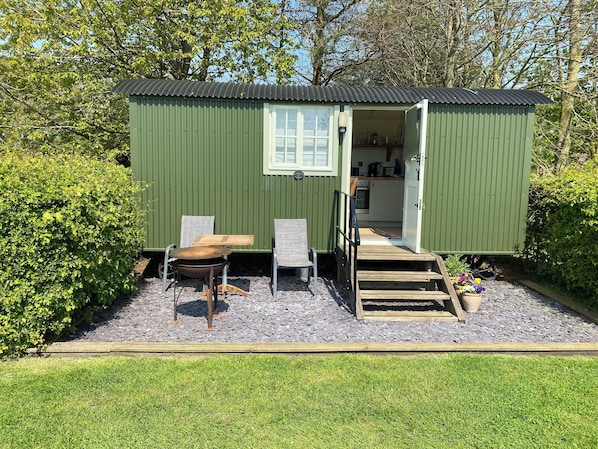 A small green metal shed with a kitchen area, two chairs, a table, and a grill outside. - Dawn Chorus Holidays (Beccles)