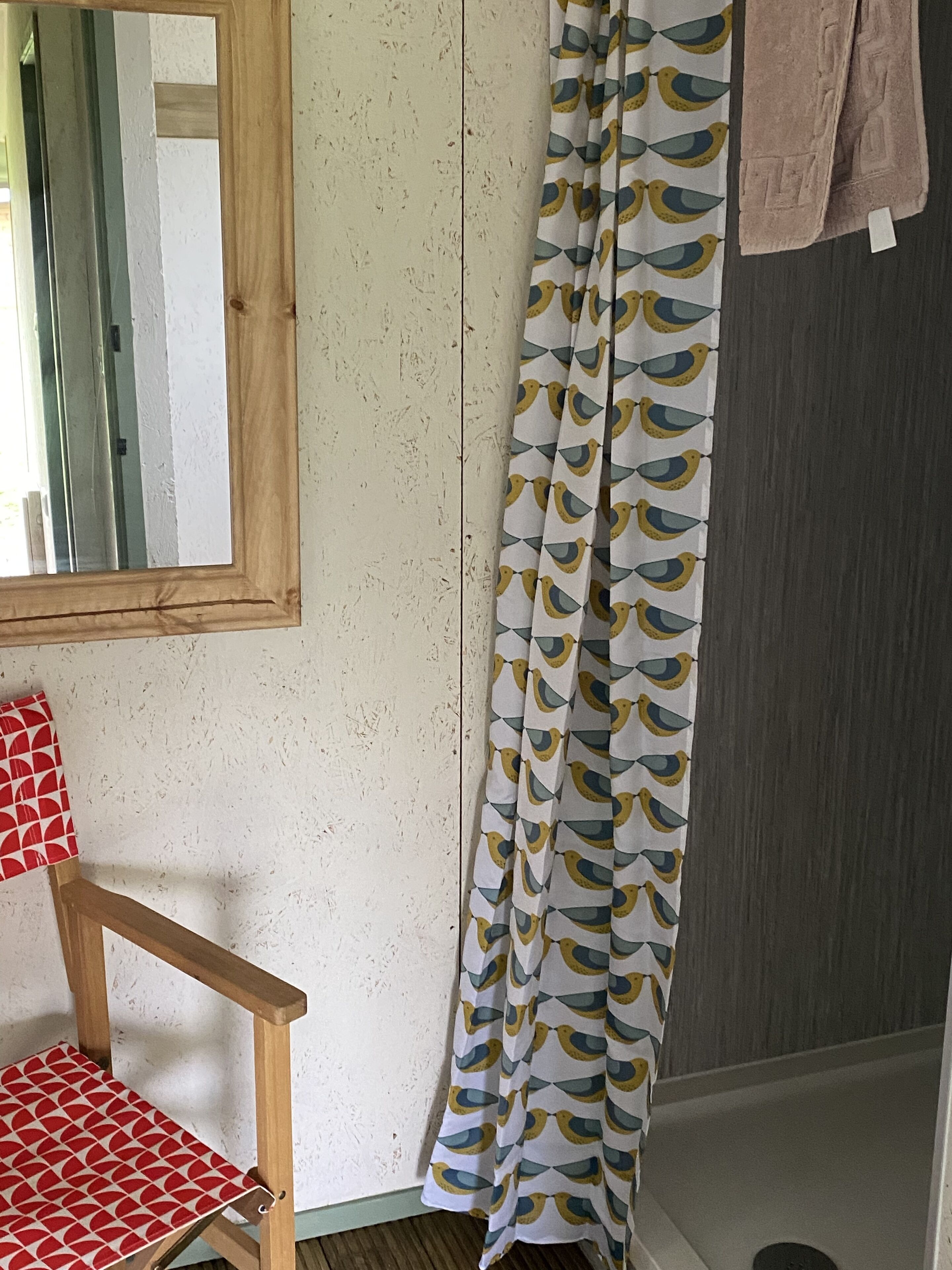 Une salle de bain avec une chaise en bois, un miroir, un rideau de douche orné de motifs d’oiseaux et un porte-serviettes.