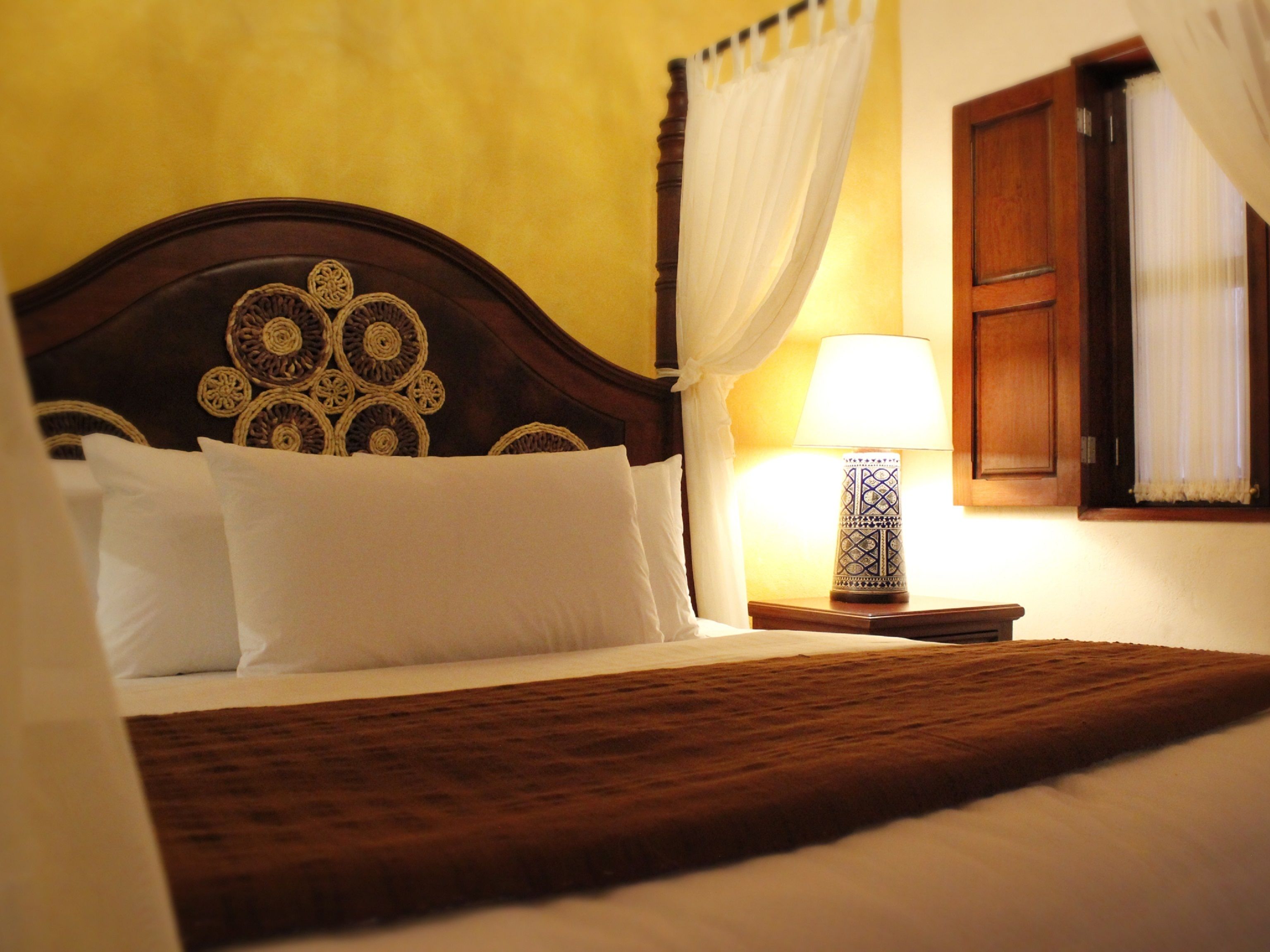 A neatly made bed with a wooden headboard, white pillows, and a brown bedspread. There is a bedside table with a lamp and a window with curtains.
