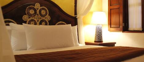 A neatly made bed with a wooden headboard, white pillows, and a brown bedspread. There is a bedside table with a lamp and a window with curtains.