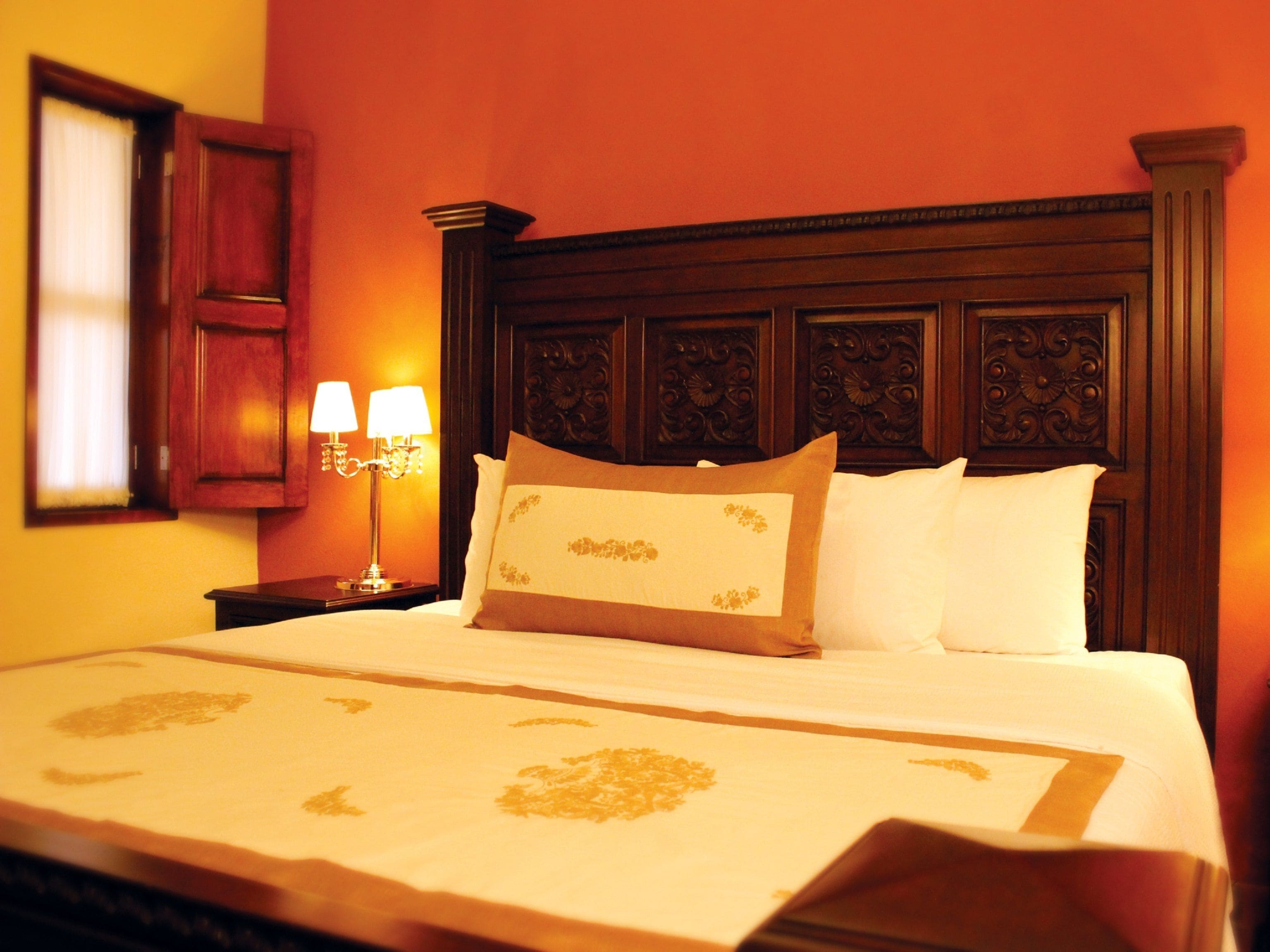 A neatly made bed with a floral bedspread, a wooden headboard with intricate carvings, and a nightstand with two lamps.
