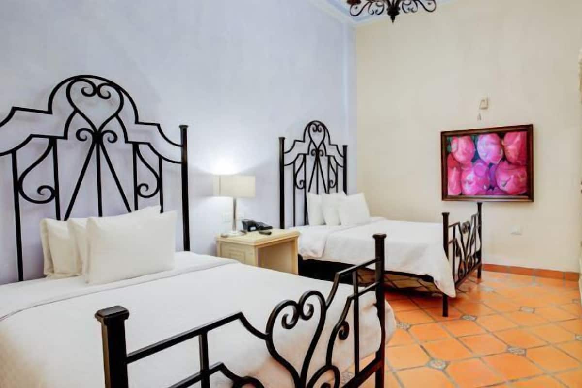 A hotel room with two beds, a desk, a chair, a TV, and a wall with a floral painting.