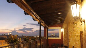 Un balcón con pared de piedra, techo de madera y vista a un paisaje urbano con una montaña al fondo.