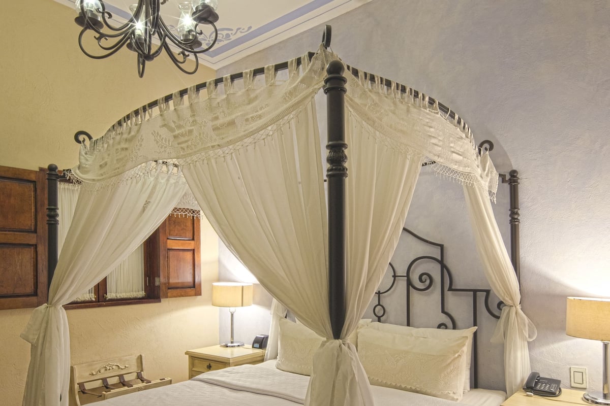 A four-poster canopy bed with a nightstand and lamp, a chandelier, and a tiled floor.