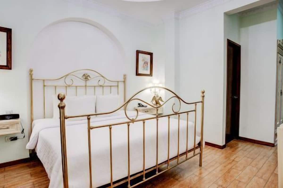 A bedroom with a bed, a nightstand, a wall-mounted shelf, and a door leading to another room.