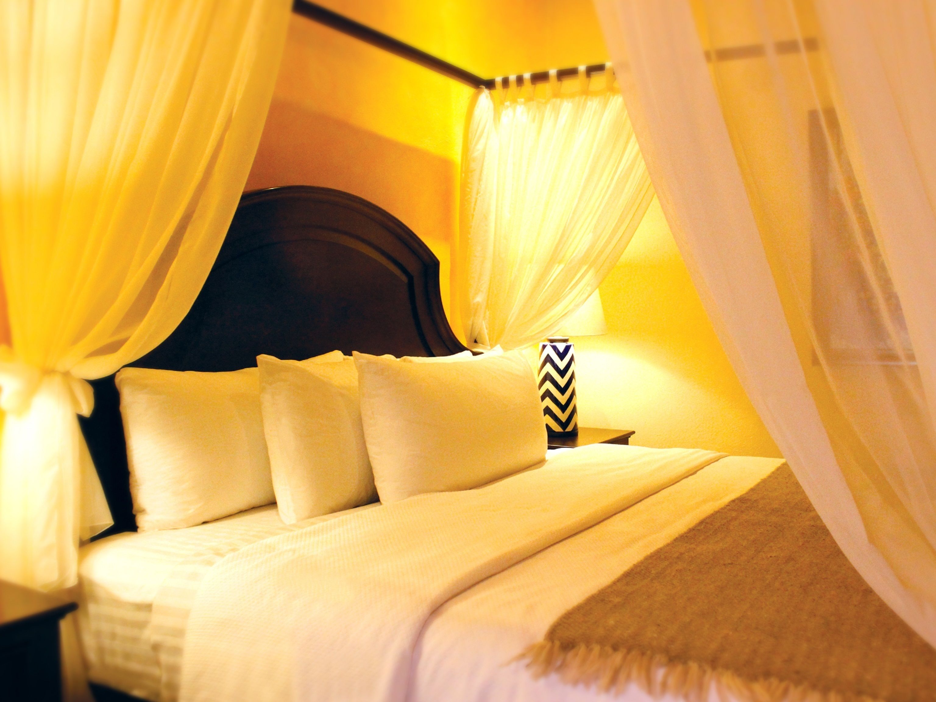 A bed with white bedding and a wooden headboard, surrounded by sheer yellow curtains.