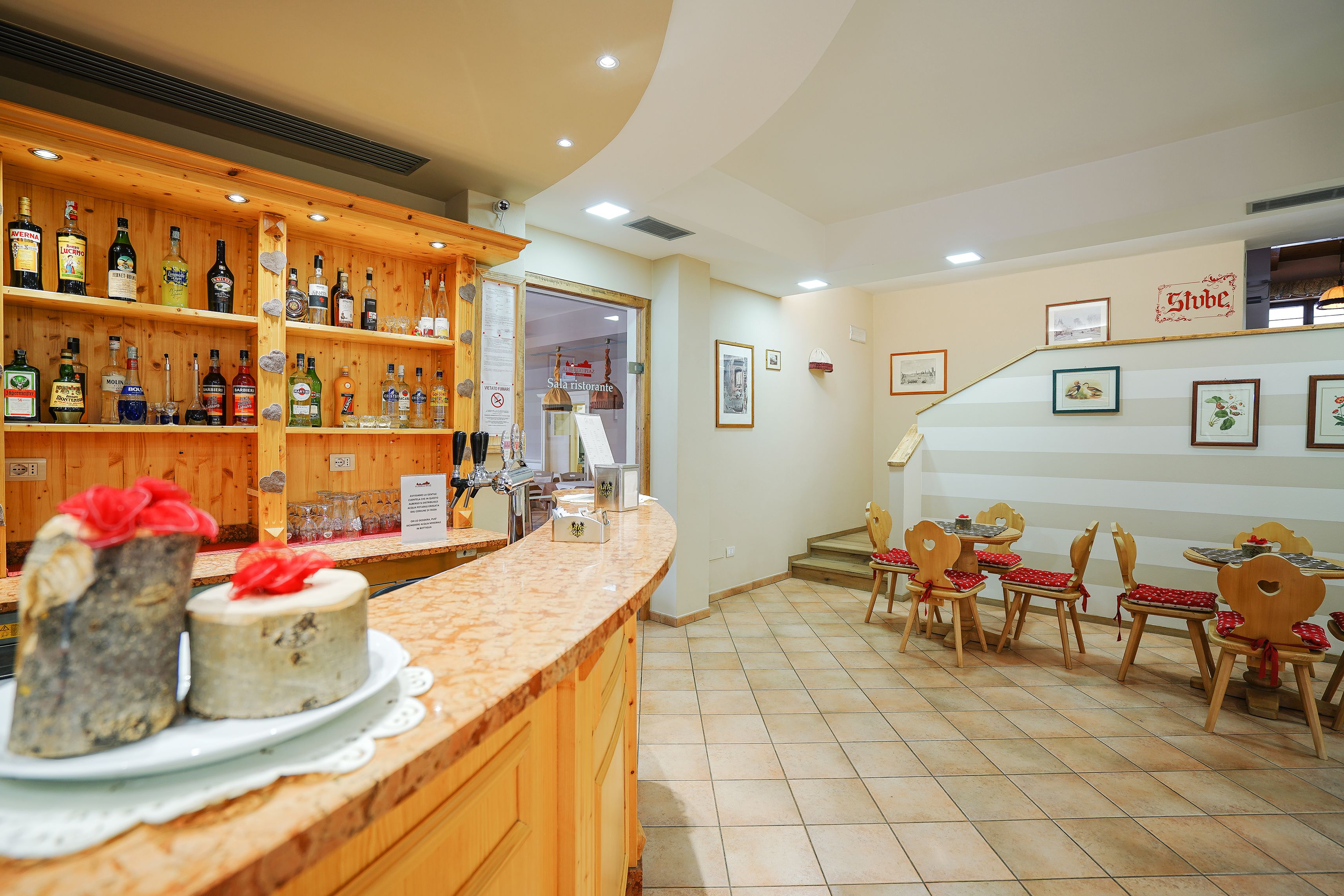 A restaurant with wooden shelves of bottles, a counter with cakes, and a dining area with chairs and tables.