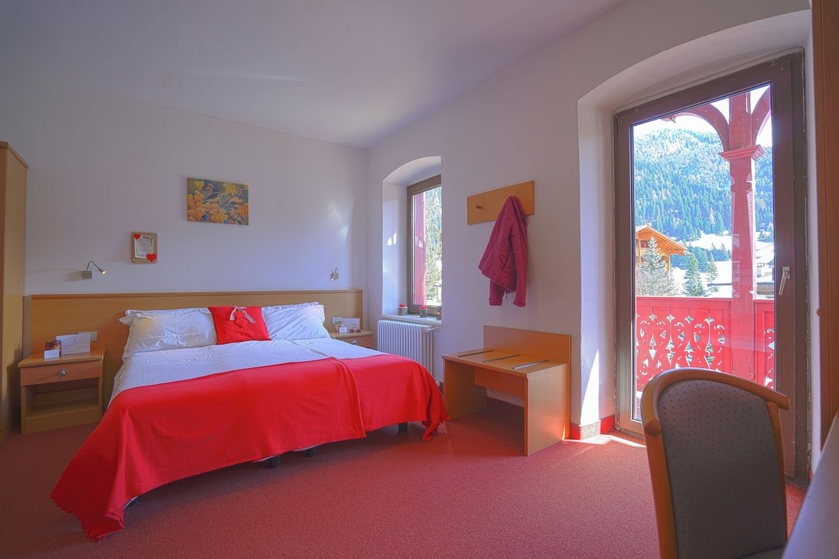 Een hotelkamer met een bed, een bureau, een stoel, een raam met uitzicht op bergen en een balkon in het rood.