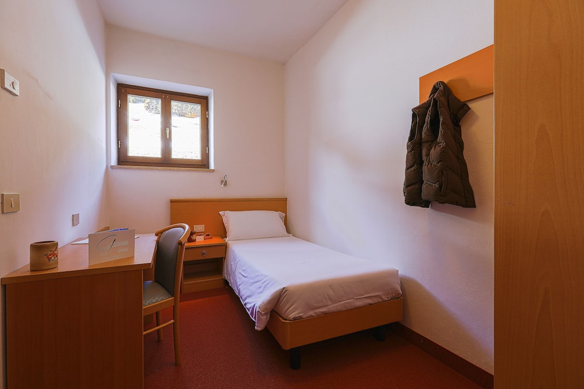 Een kleine, netjes ingerichte slaapkamer met een enkel bed, een bureau met stoel, een tafeltje en een raam.