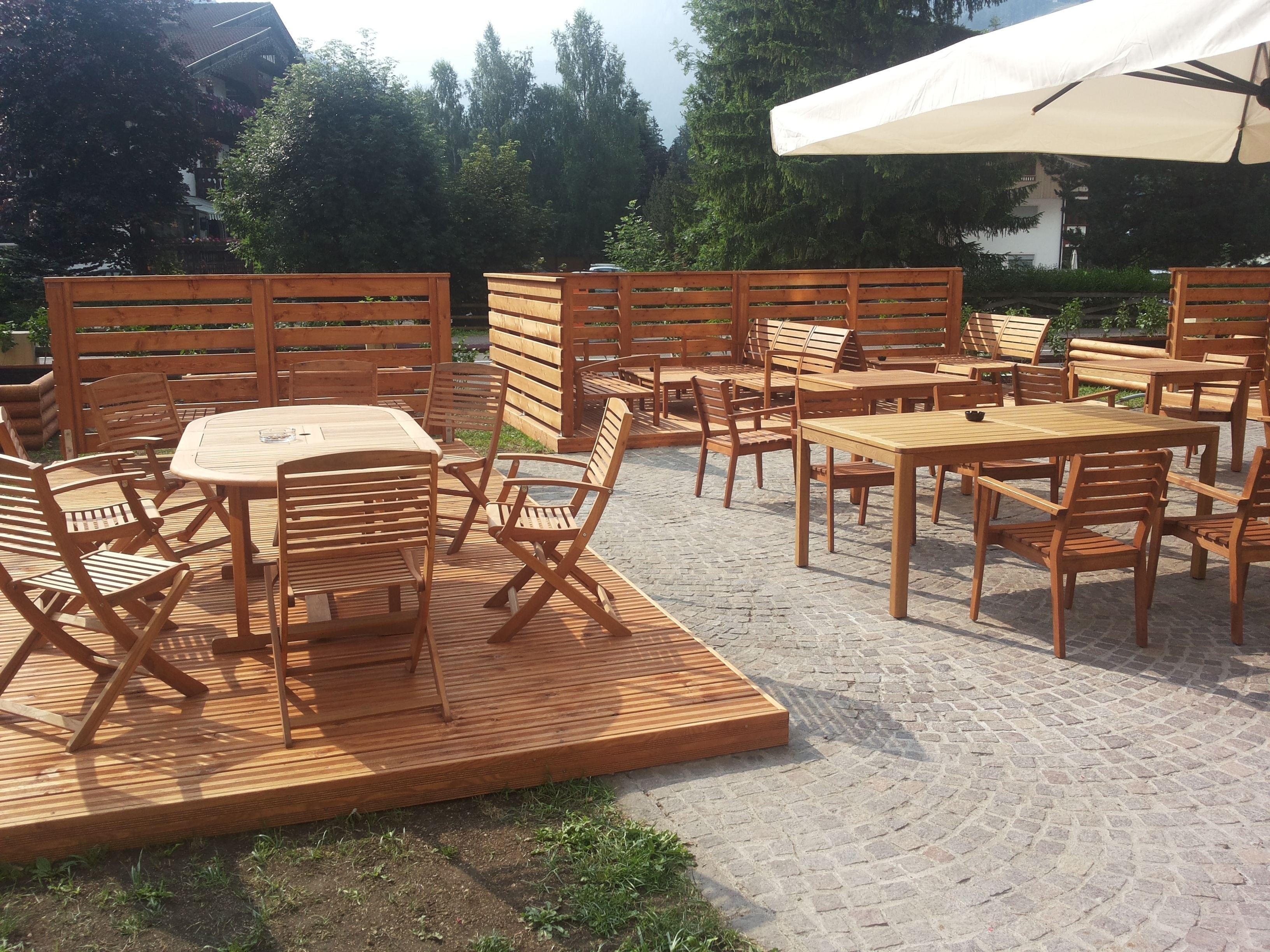 Un'area patio esterna con tavoli e sedie in legno, un ombrellone bianco e un pavimento in mattoni.