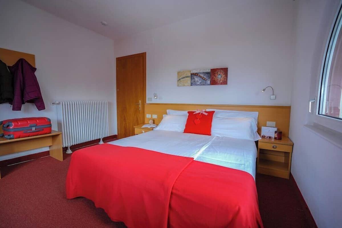 Hotelkamer met een bed, nachtkastje, rode koffer, radiator, houten deur en raam.