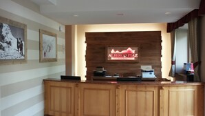 Un comptoir d’accueil avec un revêtement en bois, un téléviseur fixé au mur diffusant l’image de l’« ALBERGO PIAZ Val di Fassa », et des tableaux encadrés accrochés au mur.