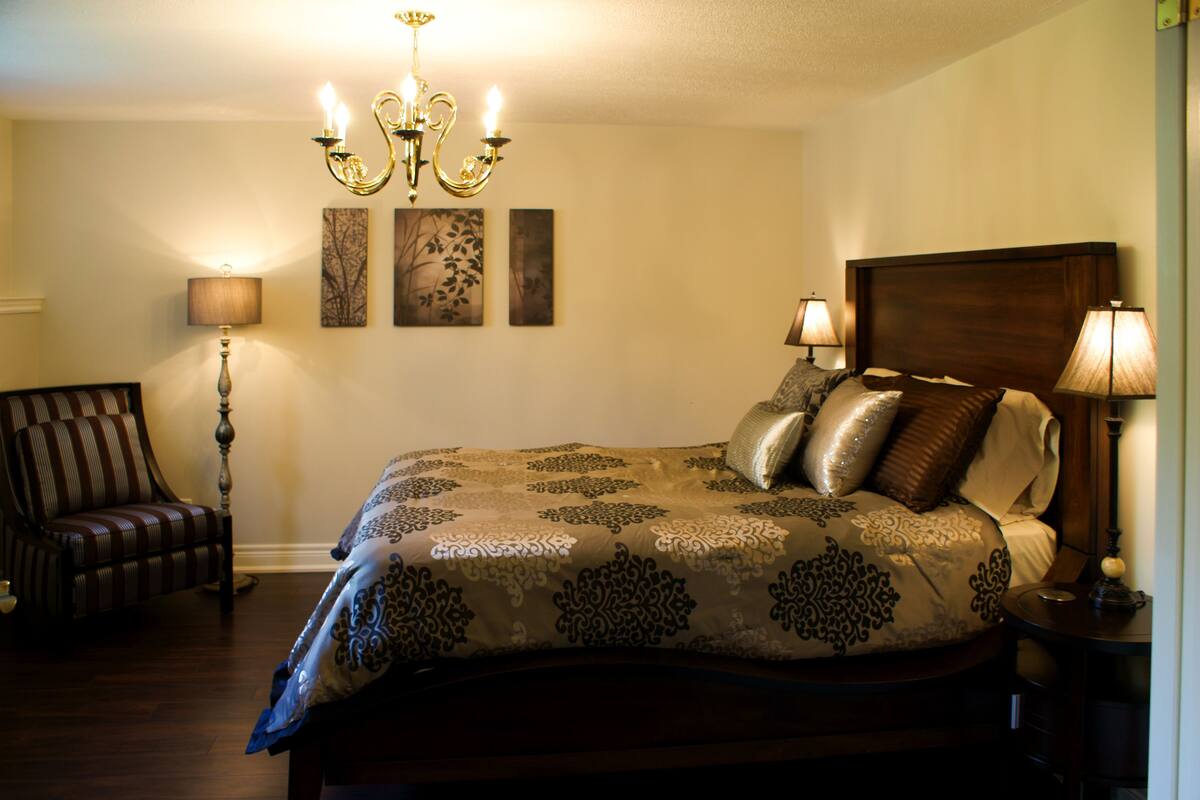 A bedroom with a large bed, two bedside lamps, a chandelier, a chair, and artwork on the wall.