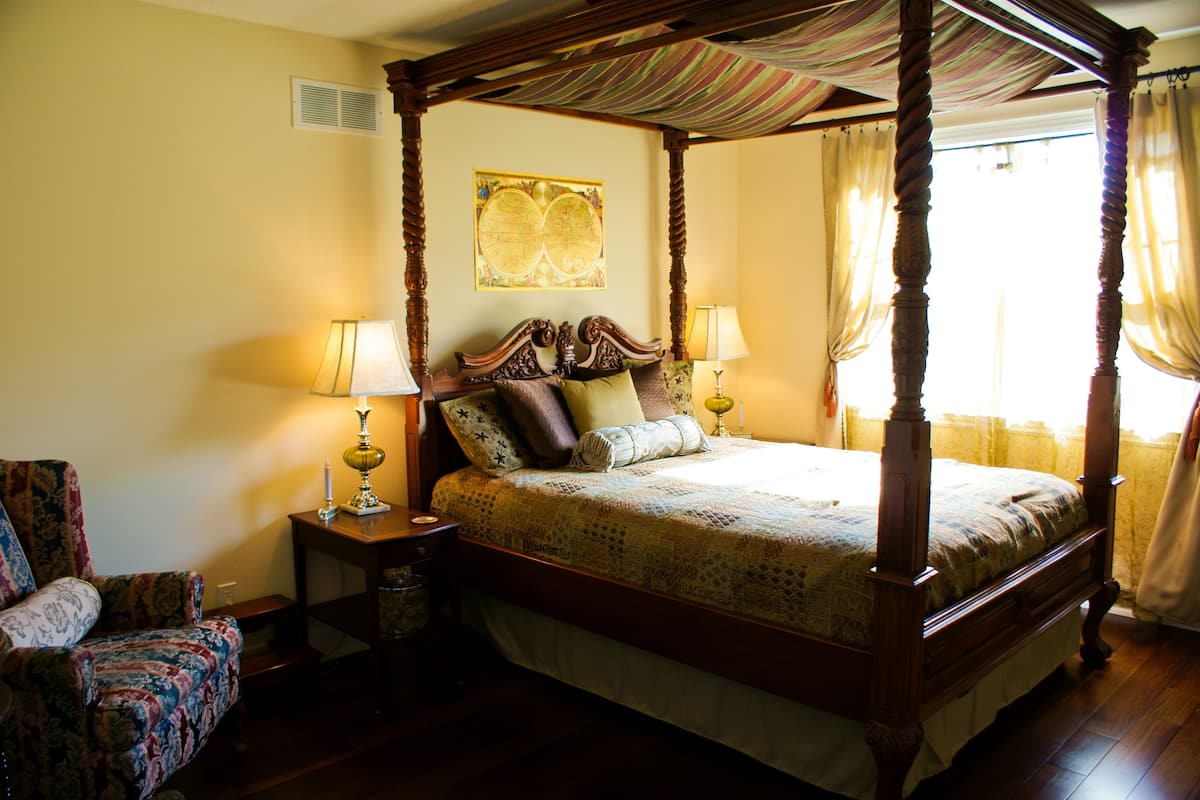 A four-poster bed with a canopy, two bedside tables with lamps, a chair, and a decorative wall piece.