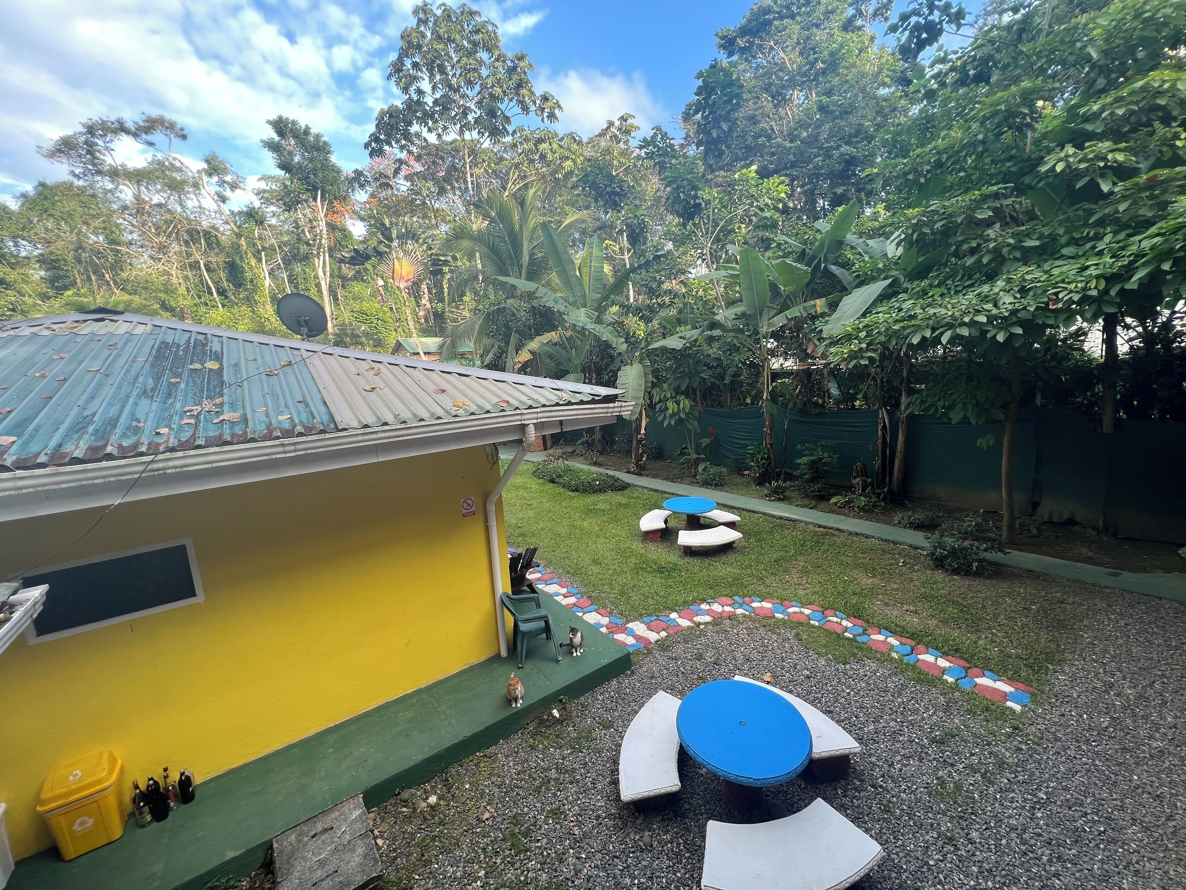 A yellow building with a metal roof, a small outdoor seating area with a blue table and white benches, and a gravel path with colorful tiles.