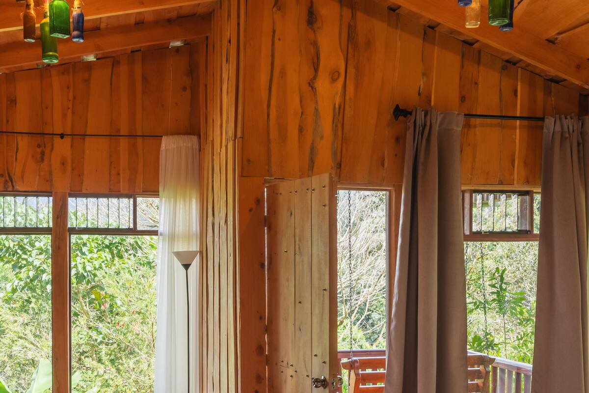 A wooden cabin with a bed, a table with chairs, and a view of greenery outside.