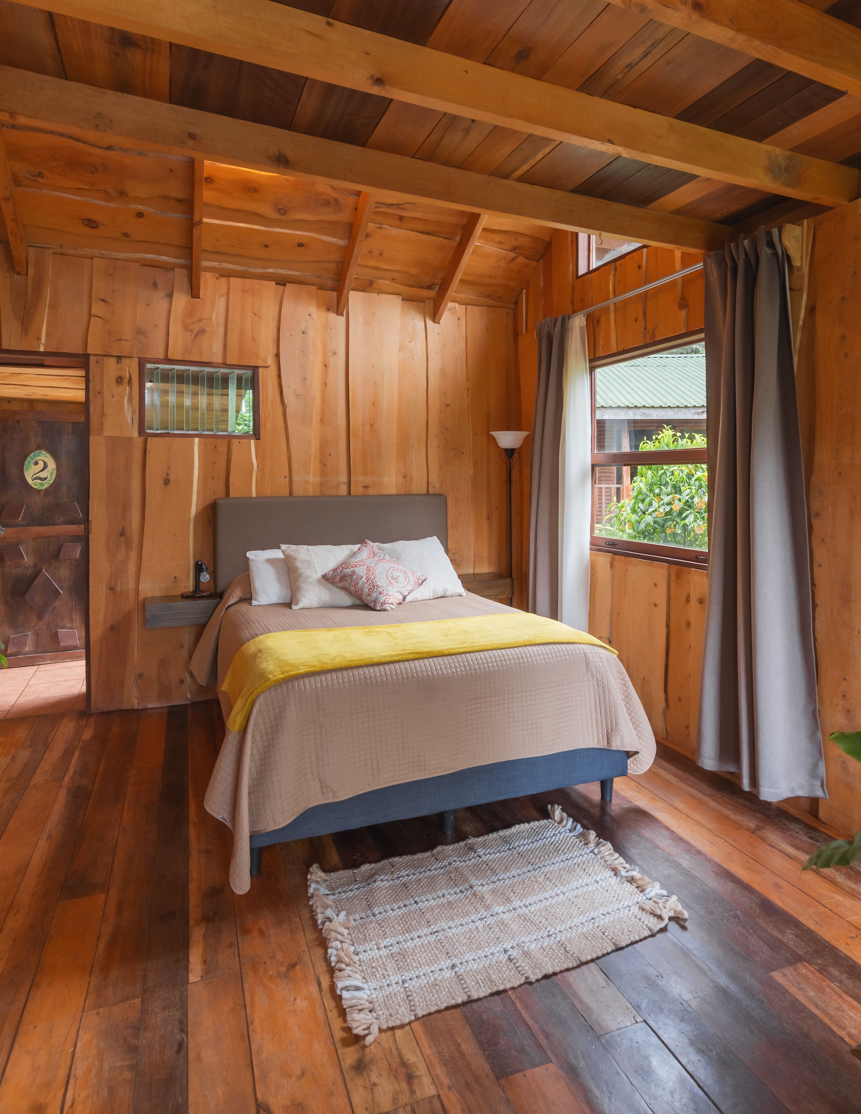 Ein Schlafzimmer einer Holzhütte mit einem Bett, einem Nachttisch, einem Fenster mit Vorhängen und einer Tür mit einer Nummer.