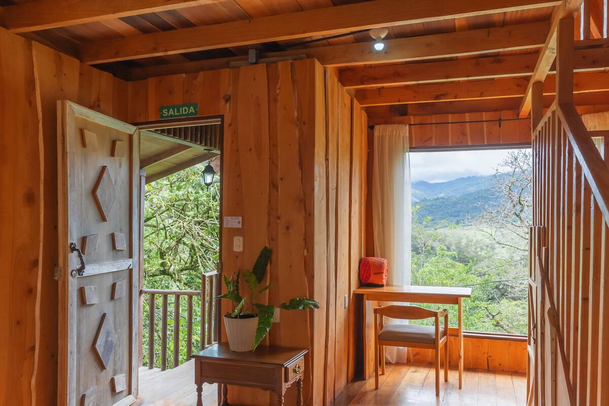 A wooden cabin room with a bed, a table with a plant, and a view of the mountains.