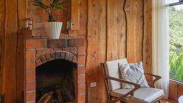 Una habitación rústica con chimenea de ladrillo, paredes de madera, una silla blanca con cojín estampado y vistas a la vegetación.