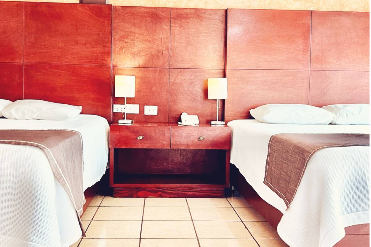 A hotel room with two beds, a wooden headboard, a nightstand with a lamp, and a small table with a phone and a cup.