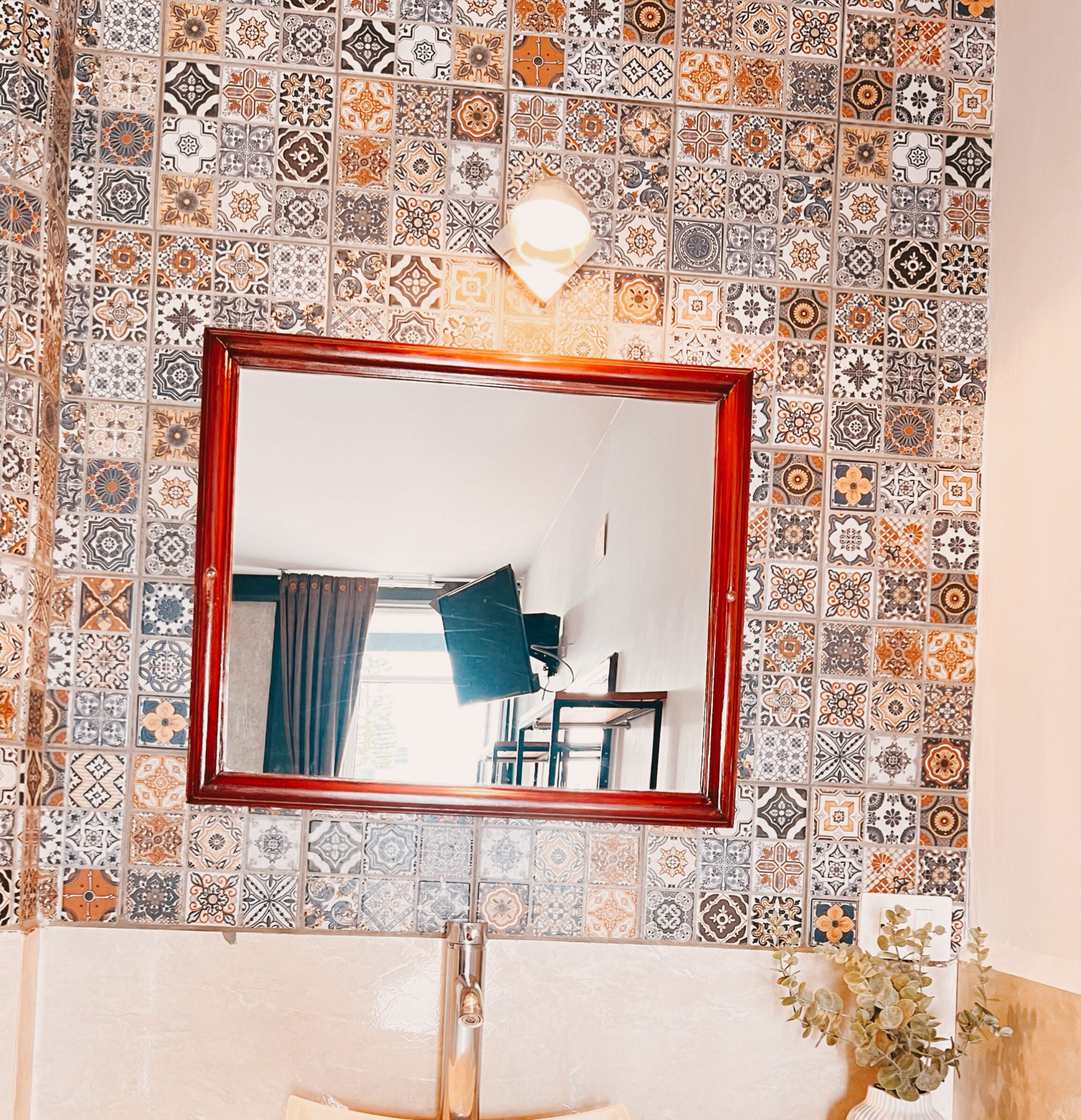 Un baño con pared de azulejos, espejo con marco de madera, lavamanos e iluminación sobre el espejo.