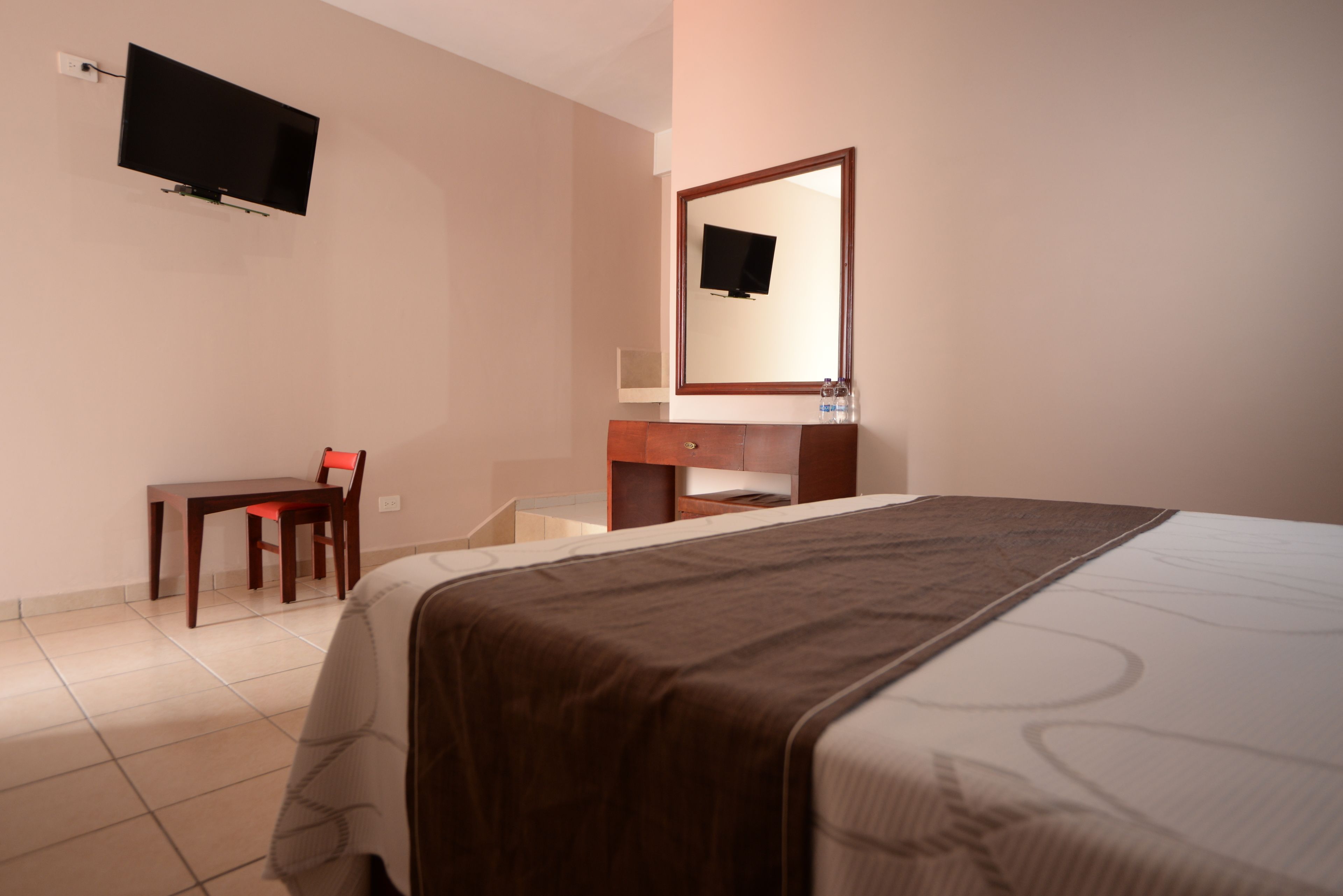 A hotel room with a bed, a desk, a chair, a TV mounted on the wall, and a mirror above the desk.
