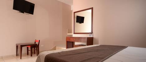 A hotel room with a bed, a desk, a chair, a TV mounted on the wall, and a mirror above the desk.