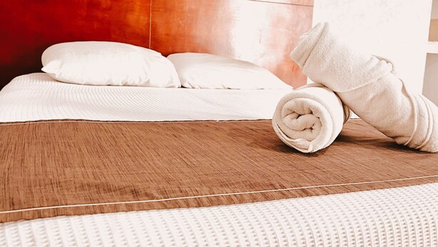 Een hotelkamer met een houten hoofdeinde, witte beddengoed en een opgerolde handdoek op het bed.