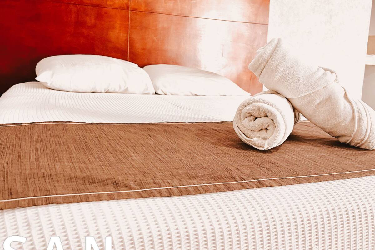 A hotel room with a wooden headboard, white bedding, and a rolled towel on the bed.