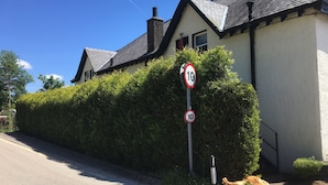 Een woonstraat met een verkeersbord voor de maximumsnelheid, een huis met een witte gevel en donker dak, en een haag langs de stoep.