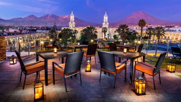 Un espacio de comedor al aire libre con mesas y sillas, faroles y vistas a un edificio histórico con dos torres y una montaña al fondo.