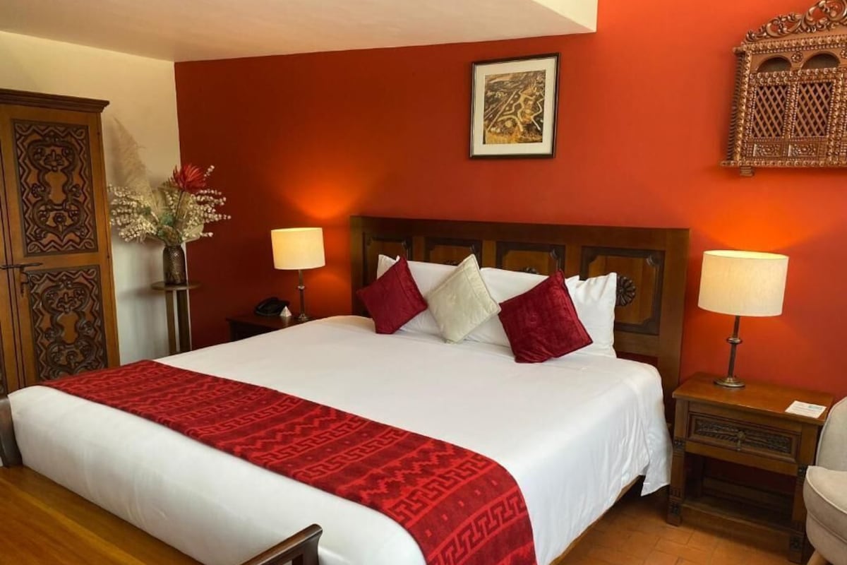 Habitación de hotel con una cama grande, dos lámparas de noche, una mesita de noche de madera, una silla de madera, un aparador decorativo y una obra de arte colgada en la pared.