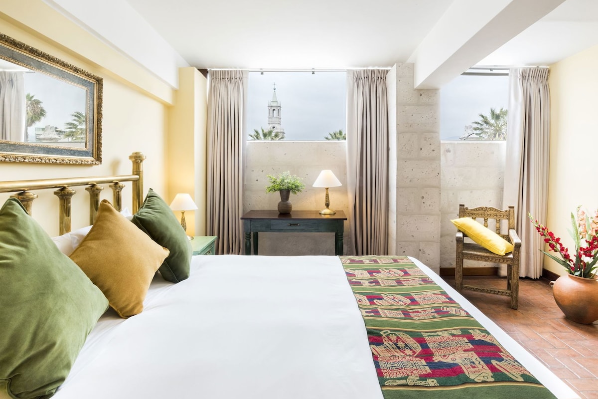 Habitación de hotel con cama, mesita de noche, silla, ventana con cortinas y vista a un edificio.