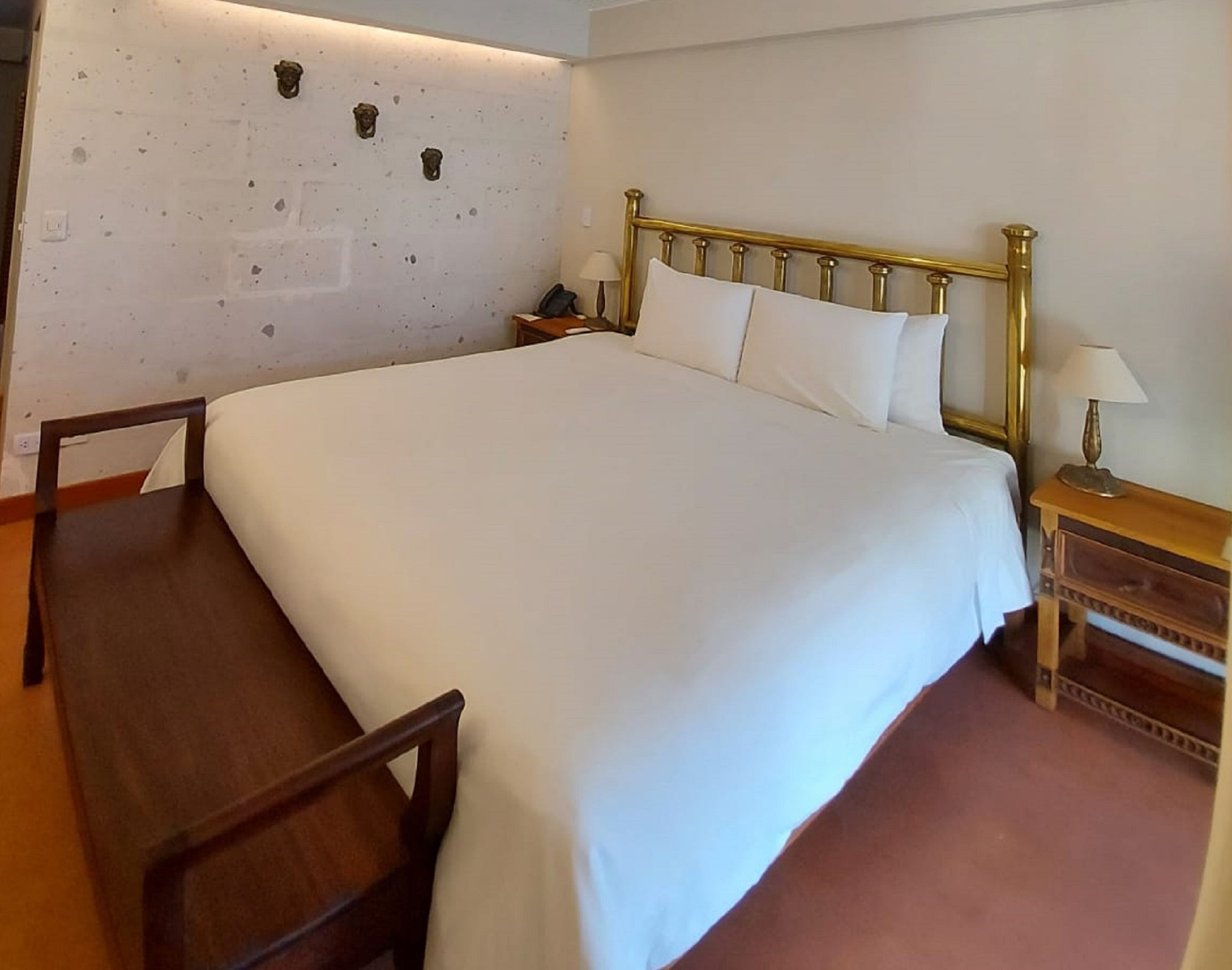 Una cama bien tendida con cabecera dorada, una silla de madera, una mesita de noche con lámpara y una pared decorada.