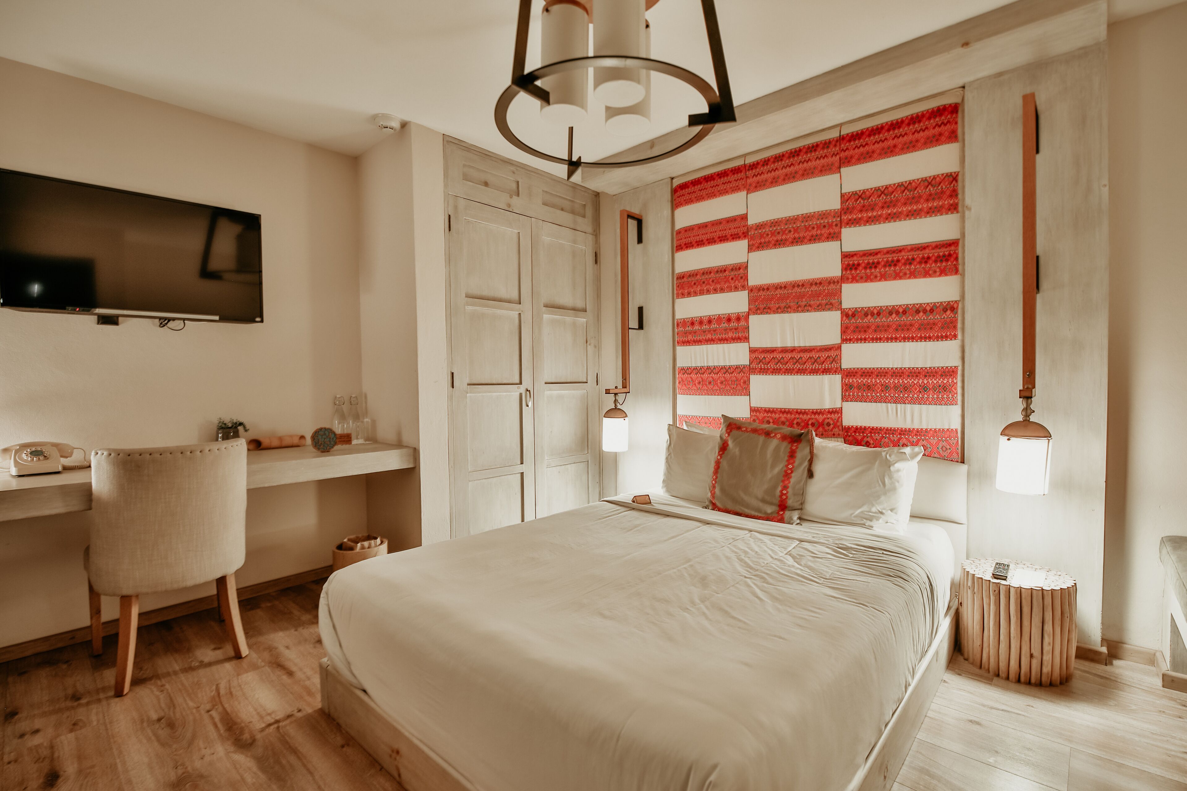 A bedroom with a large bed, a desk, a chair, a TV, and a patterned wall hanging.