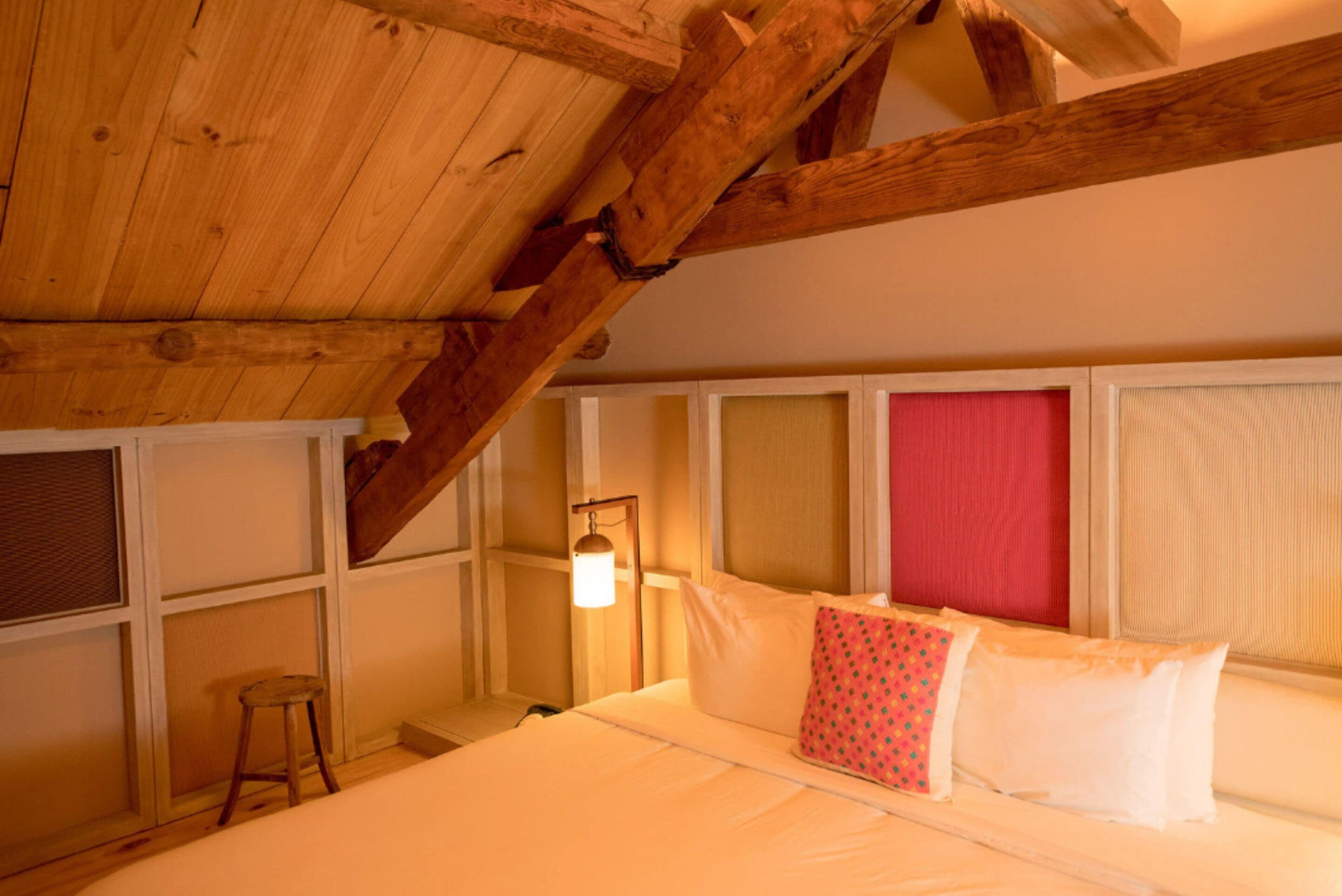 A room with a sloped wooden ceiling, a bed with white bedding and a red pillow, a small stool, and a wall-mounted lamp.