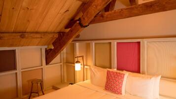 A room with a sloped wooden ceiling, a bed with white bedding and a red pillow, a small stool, and a wall-mounted lamp.