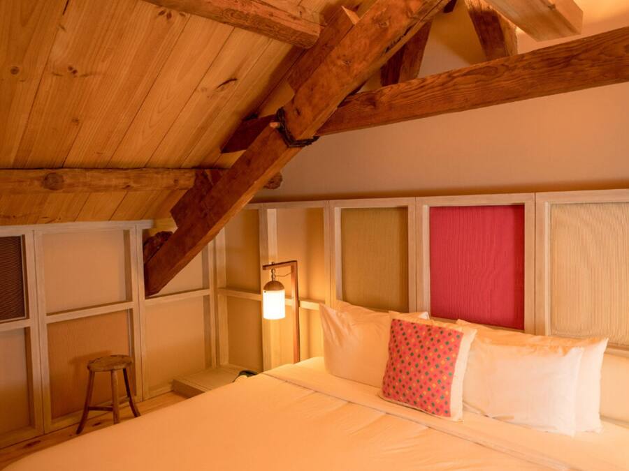 A room with a sloped wooden ceiling, a bed with white bedding and a red pillow, a small stool, and a wall-mounted lamp.