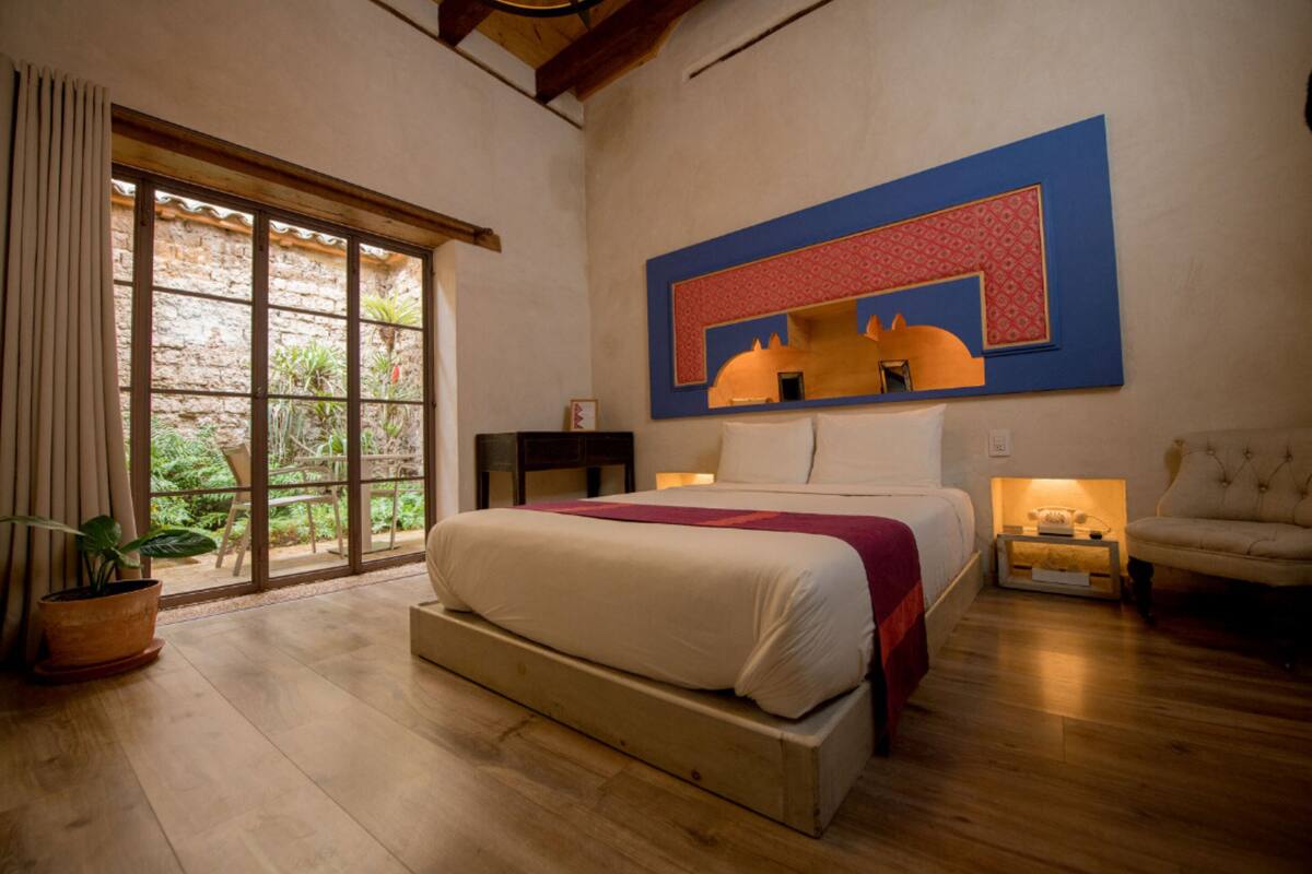 A bedroom with a large bed, a nightstand, a chair, and a view of greenery through a window.