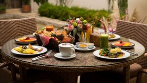 Una mesa redonda de mimbre con elementos de desayuno, incluyendo platos de comida, una cesta de pan, una taza de café y un florero con flores.