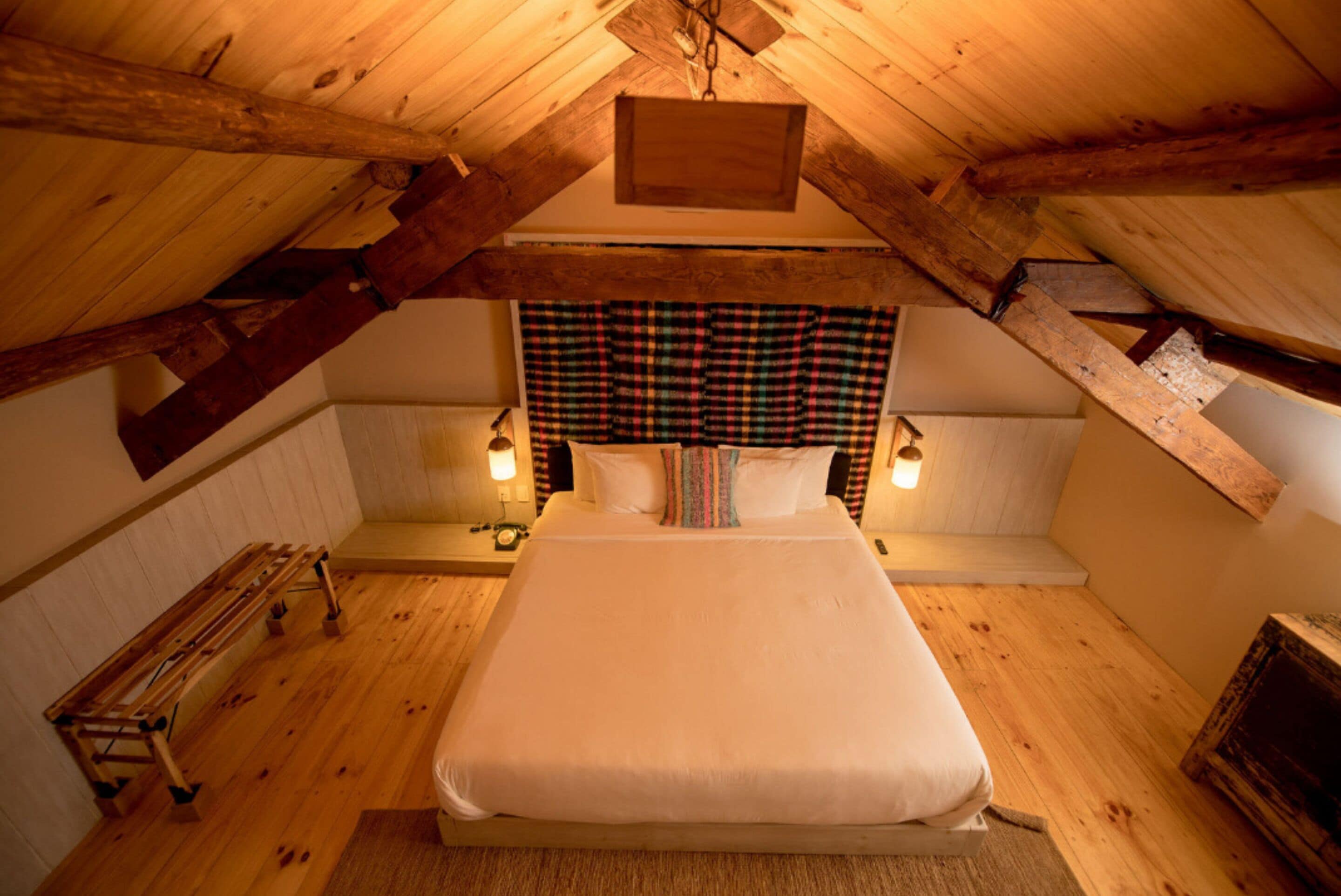 A cozy attic bedroom with a large bed, wooden beams, a plaid headboard, and two bedside lamps.