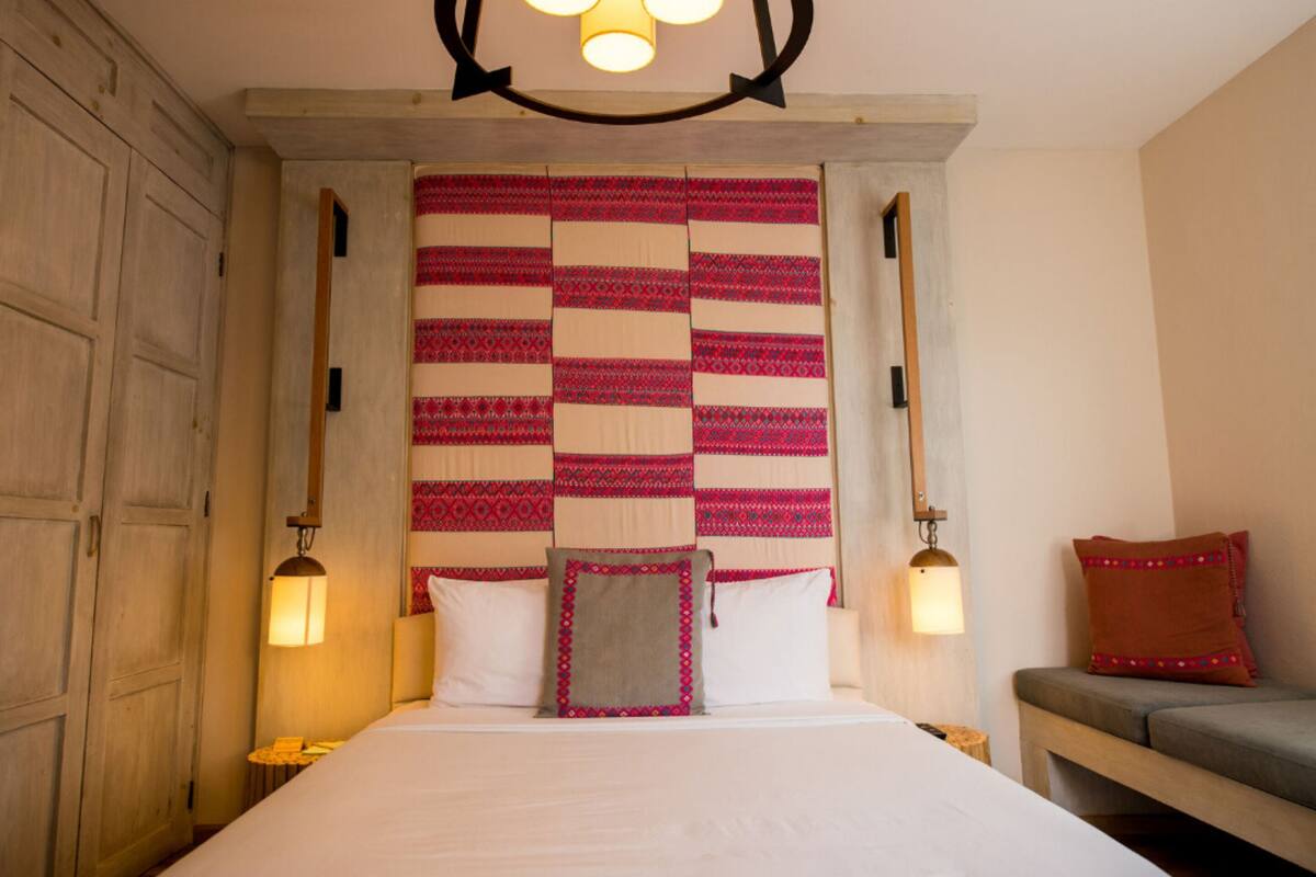 A hotel room with a large bed, two bedside lamps, a quilted headboard, a bench with a cushion, and a wooden door.