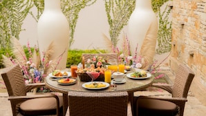 Una mesa redonda con sillas de mimbre, rodeada por un jardín con jarrones blancos y plantas.
