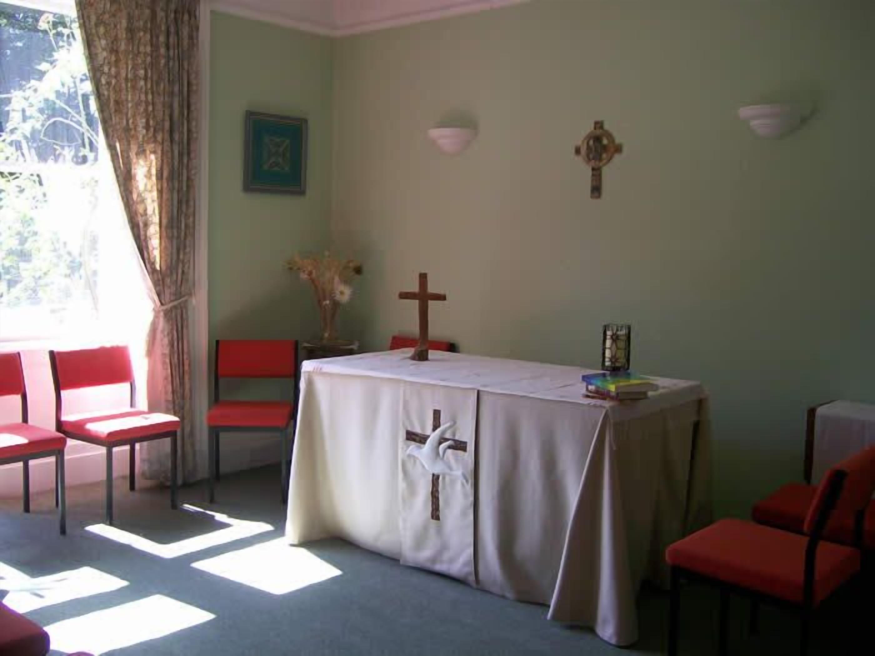 Una habitación con una mesa cubierta por un mantel blanco, sillas rojas, una cruz de madera y un cuadro en la pared.