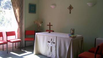 Una habitación con una mesa cubierta por un mantel blanco, sillas rojas, una cruz de madera y un cuadro en la pared.