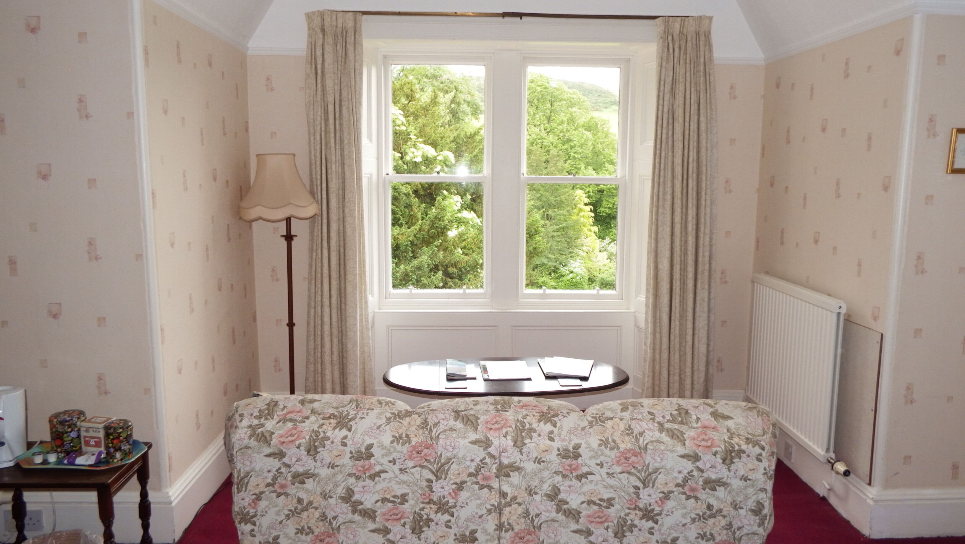 A bedroom with a bed, a window with curtains, a radiator, and a small table with a lamp.