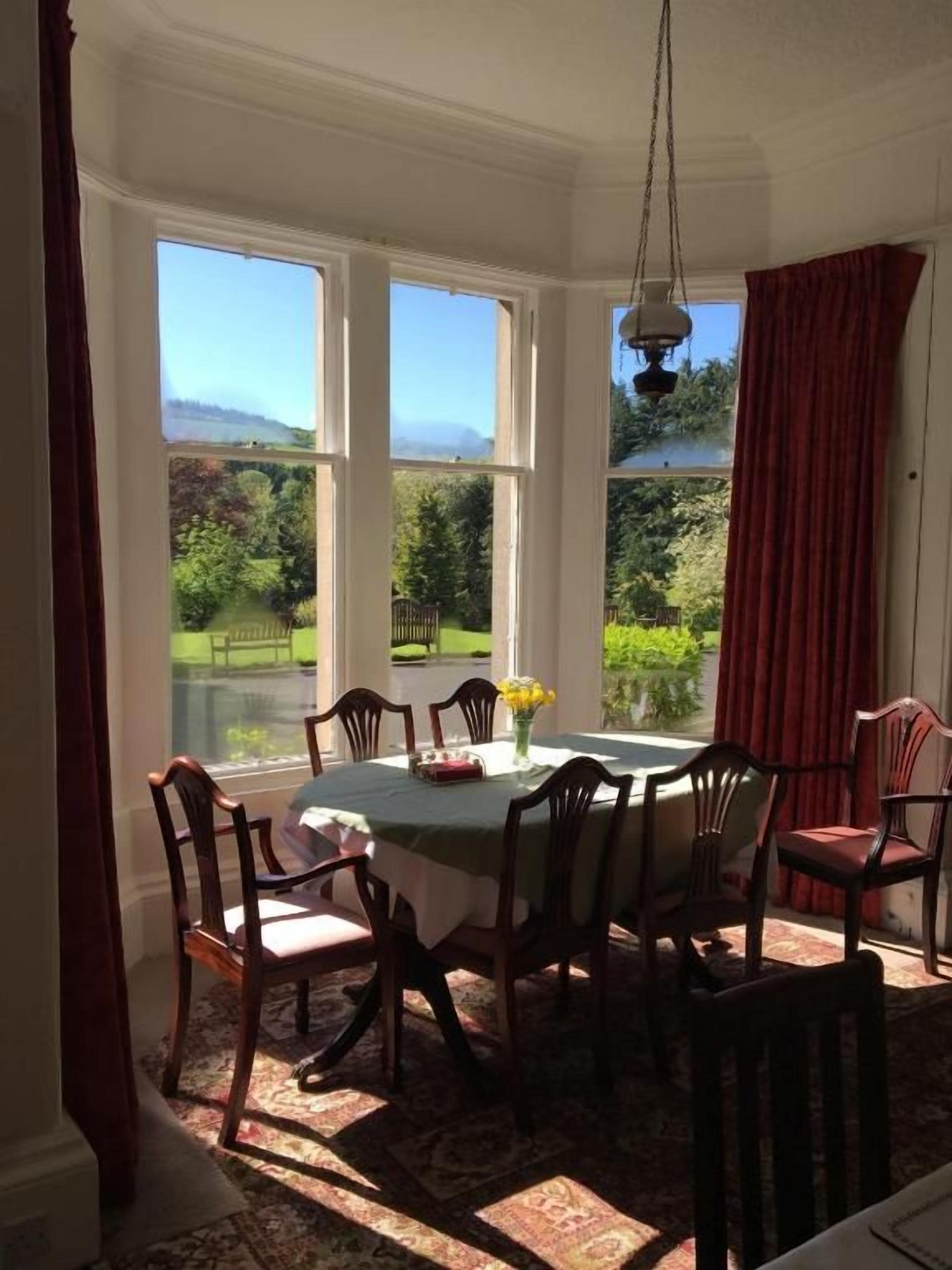 Un comedor con mesa y sillas, ventanas amplias con cortinas y vistas al exterior a zonas verdes.