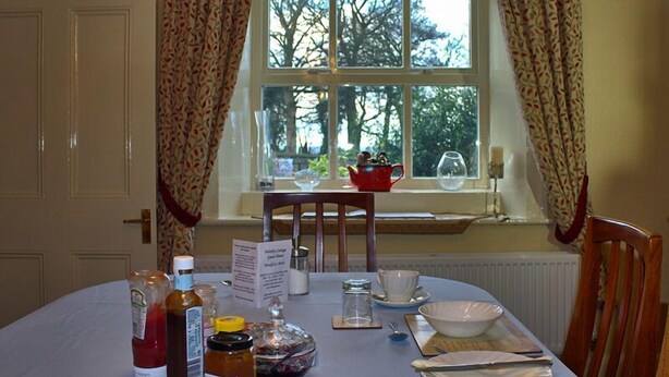 A dining table set with plates, cutlery, and condiments, with a window view of trees and a red pot on the windowsill. - Wortley Cottage Guest House (Sheffield)