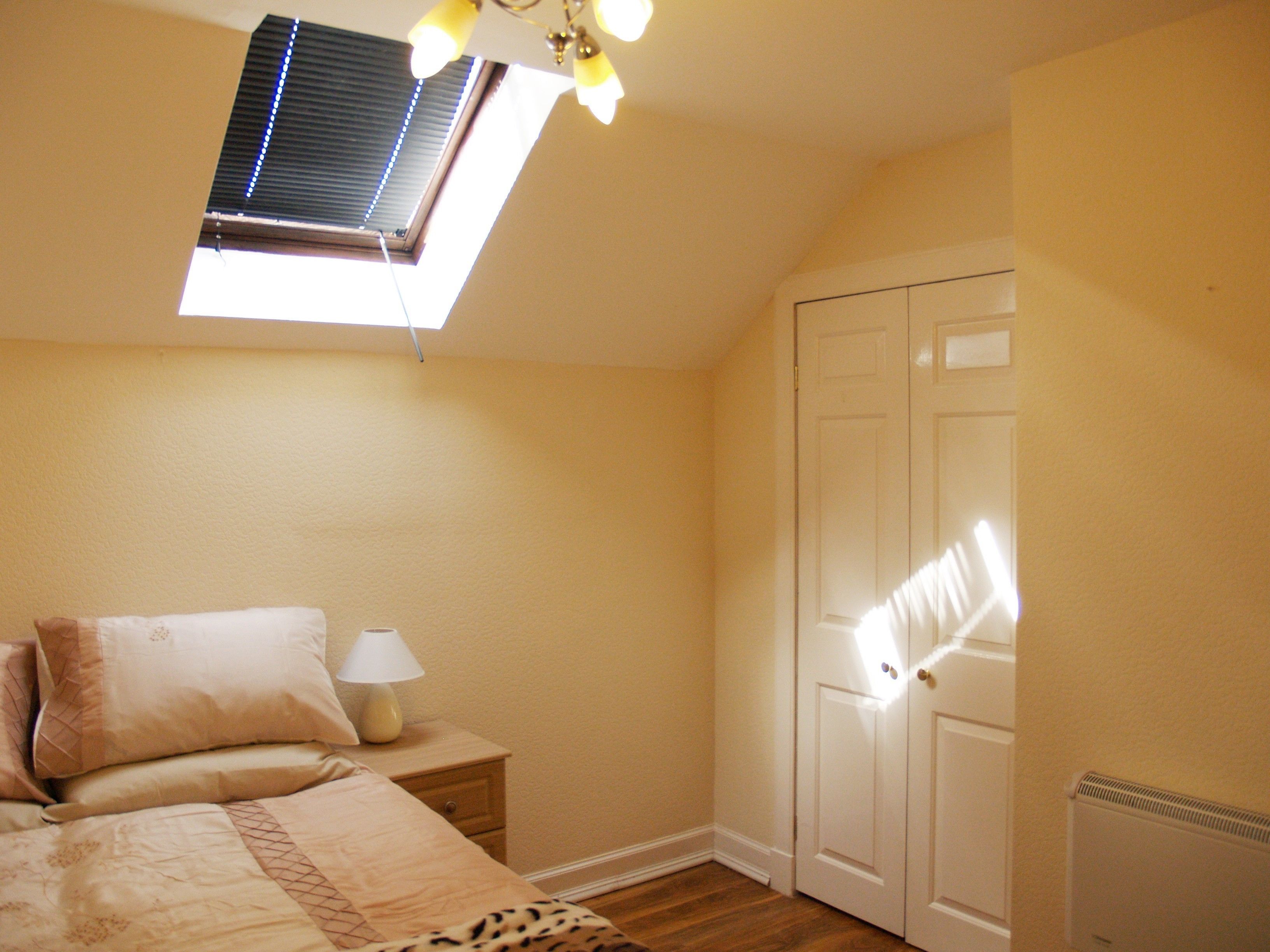 A bedroom with a skylight, a bed with a patterned bedspread, a bedside table with a lamp, and a wardrobe.