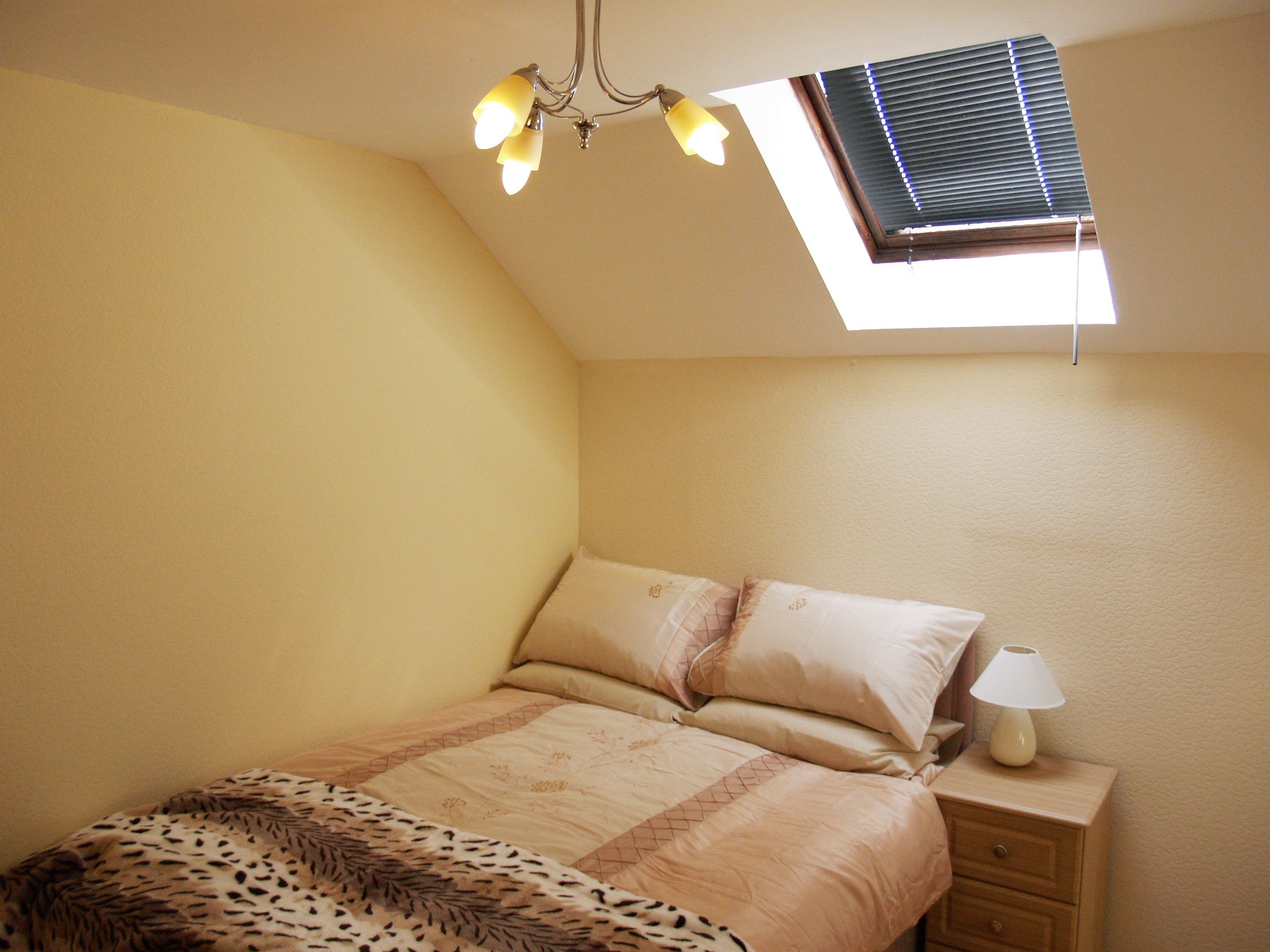 A bedroom with a sloped ceiling, a bed with patterned bedding, a nightstand with a lamp, and a window with blinds.